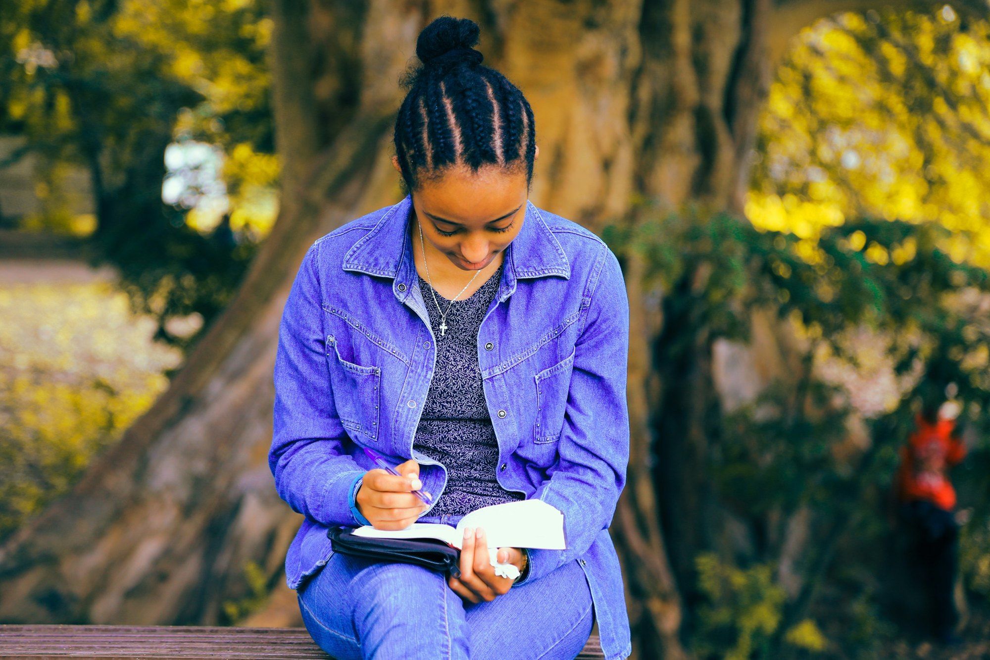 A young woman writing in a notebook outdoors under a tree.
